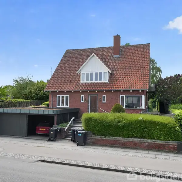 En rødstensvilla med rødt tegltag står omgivet af en velplejet hæk og have. Foran huset er en carport med en bil, og der er en flisebelagt gangsti.