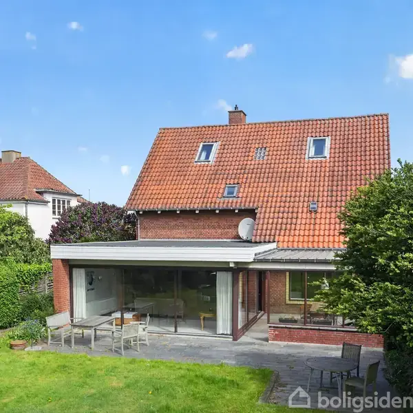 Et rødt murstenshus med en udestue, der vender ud mod en velplejet have. Der er havemøbler på terrassen, omgivet af grønne buske og træer under en klar blå himmel.