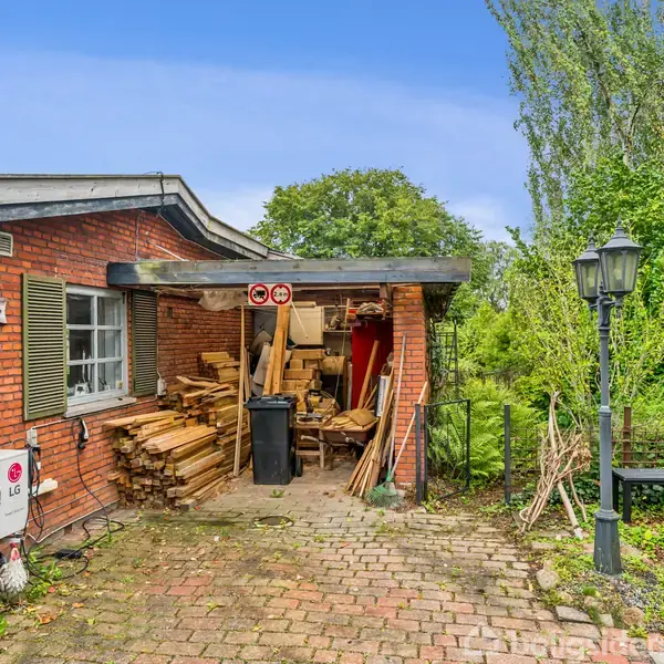 Et lille murstenshus har en overdækket opbevaring til træ, fyldt med brædder og affaldsbeholder. En luftkonditioneringsenhed er monteret på væggen. Skiltet viser teksten "Dalhøj".