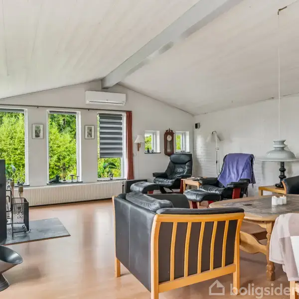 Stue med sorte sofaer og lænestole omkring et rundt træbord. Store vinduer giver udsigt til grønne træer. Væggene er lyse, og der hænger kunst på væggene.