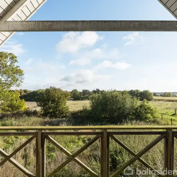 En balkon med trægelænder vender ud mod en bred udsigt over marker med spredte træer under en klar blå himmel.