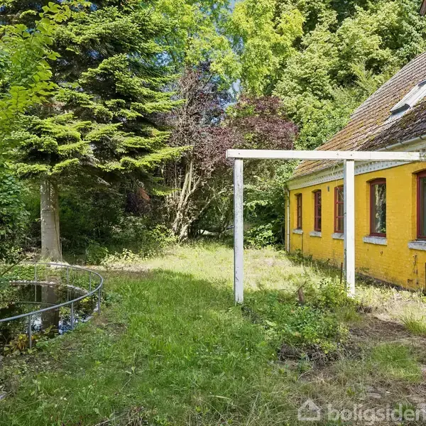 Gult hus med flere vinduer står stille ved siden af en lille afhegnet dam. Omkring huset er der frodig grøn beplantning og høje træer.