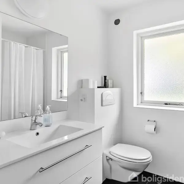 Spejl og vask i et hvidt badeværelse med minimalistisk design; toilettet er ved siden af vinduet, som slipper lys ind. Brusegardin ses delvist i baggrunden.