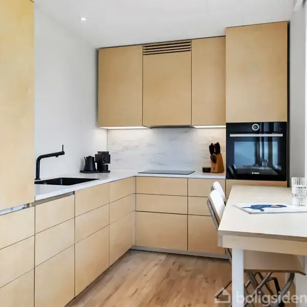 Et moderne køkken med lyse træskabslåger, en indbygget ovn, og en sort vandhane ved en hvid bordplade. Der er et lille siddebord ved vinduet med en stol.