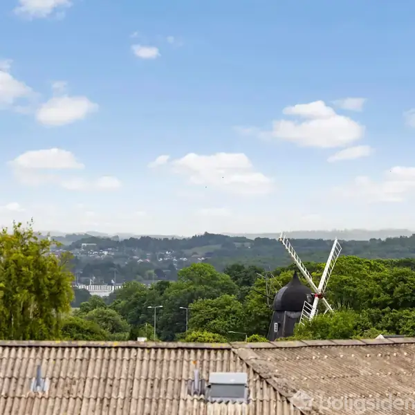 En mølle står stille omgivet af grønne træer, set over hustage. Baggrunden viser en klar blå himmel med spredte skyer.