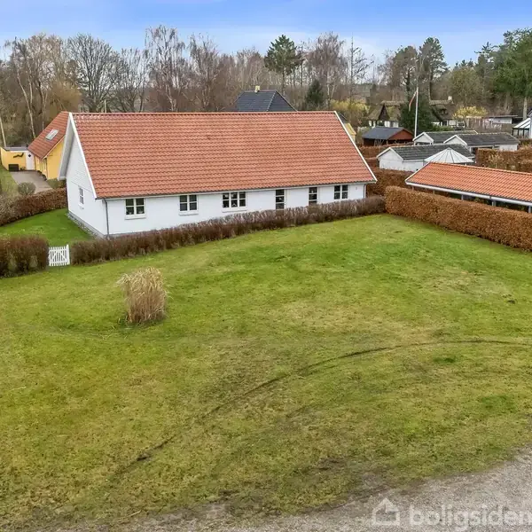 Hvidt hus med rødt tag står stille omgivet af en stor græsplæne og hække. Flere huse og træer ligger i baggrunden under en klar himmel.