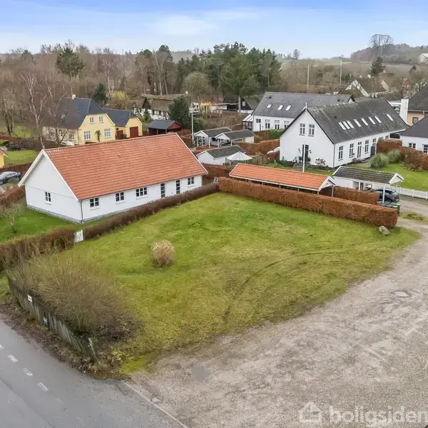 En hvid bygning med rødt tag står stille i et åbent grønt område omgivet af lave buske. Omkring ses flere huse og en parkeret bil på en grusvej.