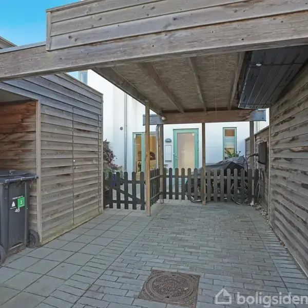 Carport med åbent trætag og flisebelagt gulv, beliggende ved et husindgangsområde. En skraldespand står ved siden af et postkasseområde med nummeret "46". En lav hegn adskiller områderne.