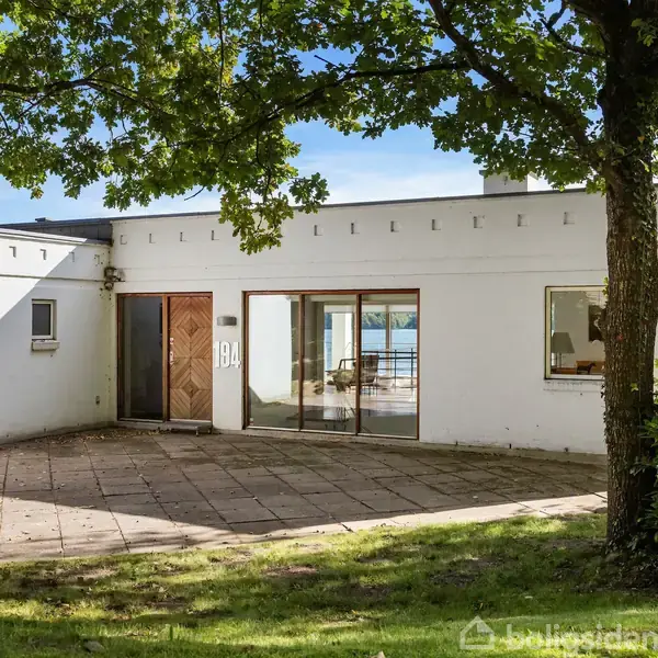 En hvid modernistisk villa med store vinduer og trædør, står stille på en flisebelagt terrasse omkranset af græs. Baggrunden viser en sø og grønne træer. Husnummeret "19A" ses ved døren.