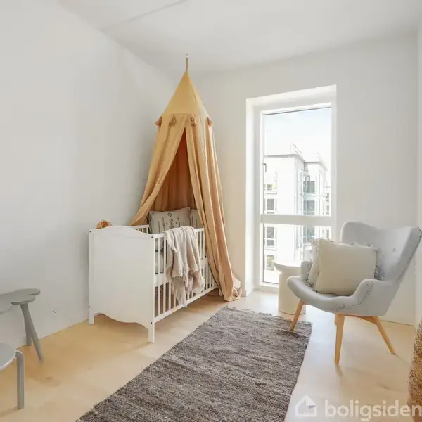 Værelsesindretning med hvid tremmeseng under gul baldakin. Gråt tæppe ligger på gulvet. Lysegråt loungesæt ved vinduet. Små grå borde med legetøj i hjørnet. Hvidmalede vægge.