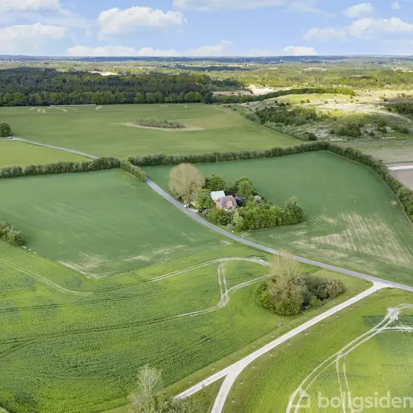 Et hus omgivet af grønne marker ligger ved en vej, med skove og marker der strækker sig mod horisonten under en blå himmel med hvide skyer.