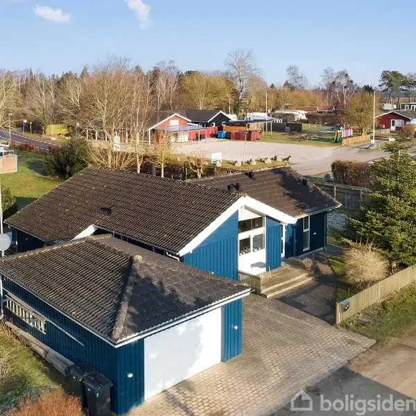 Blåt hus med sort tag, stående på flisebelagt indkørsel, omgivet af buske og træer i et nabolag med andre huse og veje.