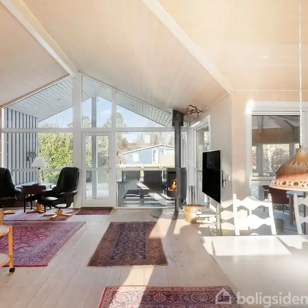 Rummelig stue med højt til loftet, store vinduer, og gulvtæpper. Sorte lænestole står ved borde. Et spisebord med lamper hænger i nærheden. Udsigt til grønne træer udenfor.