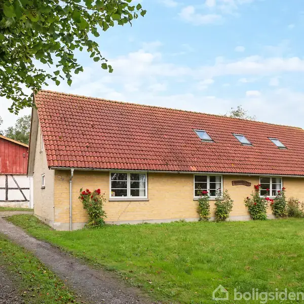 Et gult murstenshus med rødt tegltag står i en grøn have omkranset af en grussti. Til venstre ses en rød lade, og himlen er klar med skyer.