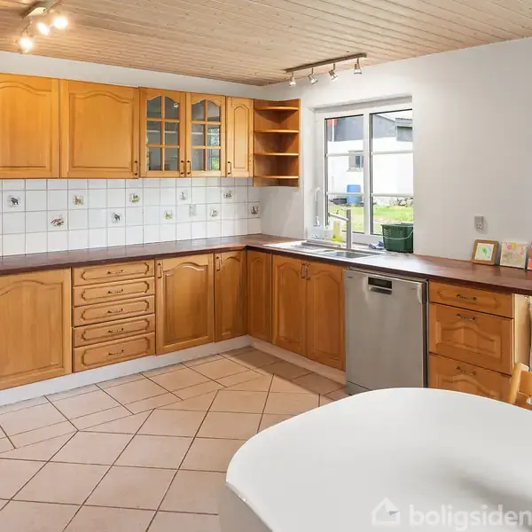 Køkken med træskabe, flisevæg og opvaskemaskine, udenfor et vindue. Rummets lyse fliser og træloft skaber en varm atmosfære.