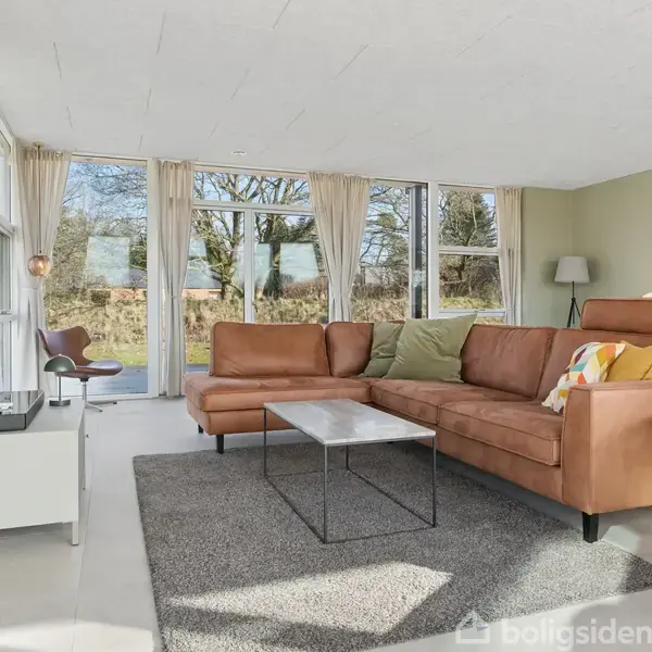 Brun sofa står i lyse stue med glasvægge og udsigt til træer. Gråt tæppe og sofabord supplerer den moderne indretning. Et stort fladskærms-tv hænger på væggen.
