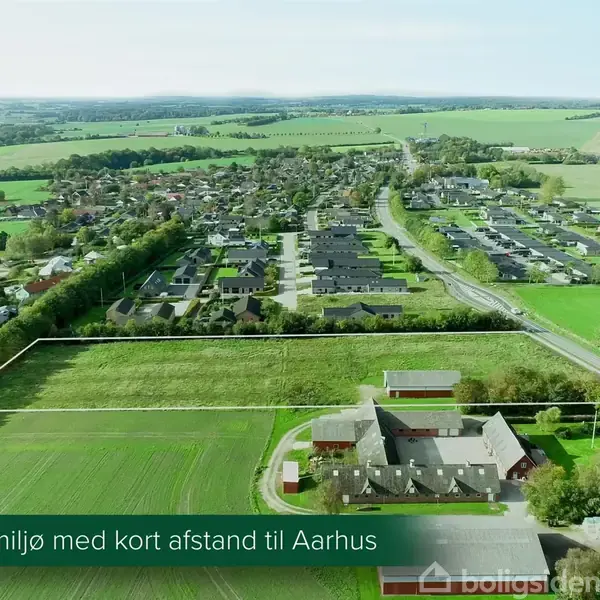 En åben græsmark med markerede grænser ligger i forgrunden, mens en landsby strækker sig i baggrunden under en klar himmel. Tekst: "Attraktivt nærmiljø med kort afstand til Aarhus".