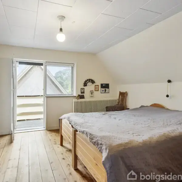 Soveværelse med dobbeltseng og sengebord, åbent til en altandør. Trægulv og hvidt loft med en enkelt lampe. Skab langs venstre væg og stol ved vinduet.