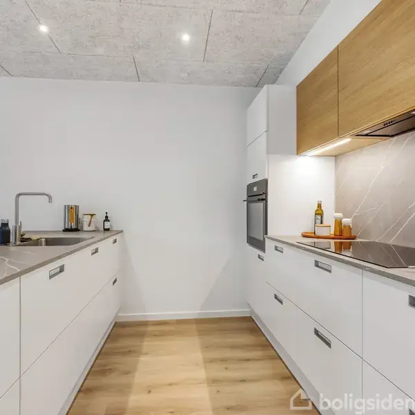 Et moderne køkken med hvide skabe er udstyret med en integreret ovn. En køkkenvask og diverse køkkenredskaber er på en marmorbordplade. Loftet har indbyggede spotlights.