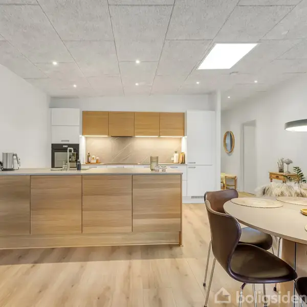 Køkken med træelementer, hvidevarer og lys bordplade. Spisebord med stole i forgrunden, omgivet af moderne indretning. Loftet har indbygget belysning. Vægpyntegenstande ses i baggrunden.