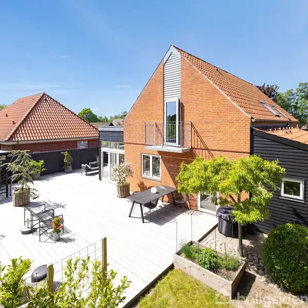 Rødt murstenshus med hvide vinduer står i solskin. Terrasse med møbler og planter omkranser huset. Græsplæne og træer i baggrunden skaber en hyggelig haveatmosfære.