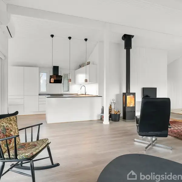 En moderne stue med en sort brændeovn står tæt på et åbent køkken, der har hvide skabe og hængende lamper. En gyngestol og lænestol er placeret på trægulvet.