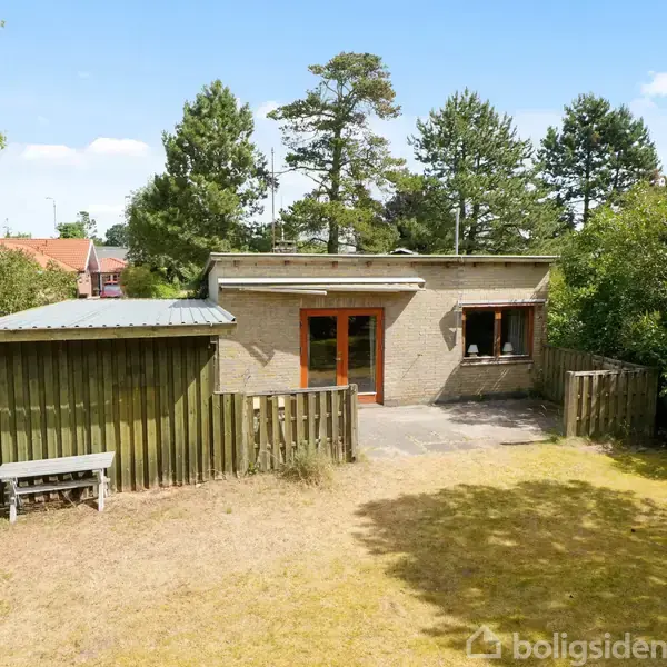 Et lille hus med fladt tag og store trædøre åbner ud til en terrasse. Omgivet af en frodig have med træer og buske.