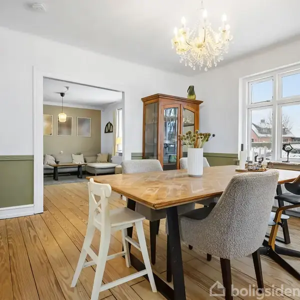 Spisebord med stole står i lyst rum med trægulv, lys lysekrone hænger over. I baggrunden en stue med sofaer og et vitrineskab ved væggen. Store vinduer giver udkig til haven.