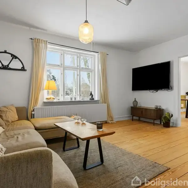 En lys stue med en beige sofa står ved et træbord. En stor fladskærm hænger på væggen. Rummet har trægulv, vindue med gardiner og en dør til et spisestueområde.