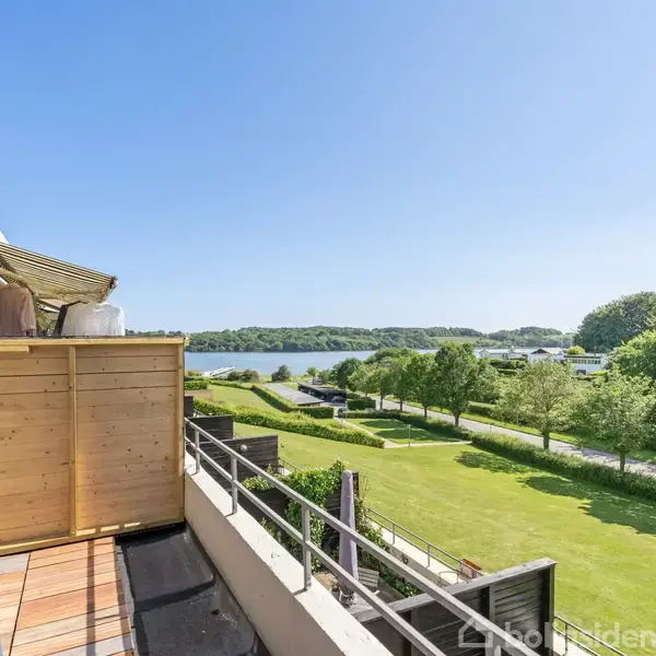 En altan med en trævæg vender ud mod en sø og grønne områder på en solrig dag. Græsplæner og træer strækker sig mod vandet under en klar blå himmel.