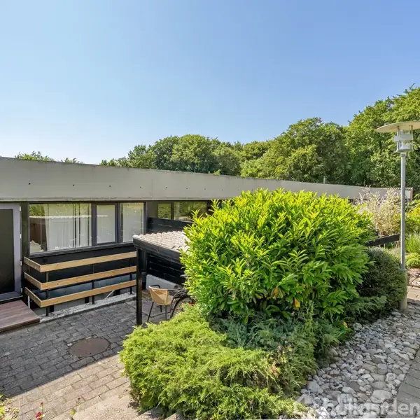 Rækkehus med fladt tag står ved siden af en brolagt gangsti. Foran huset er der en velplejet have med buske og græs. Bagerst ses træer og en blå himmel.