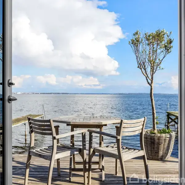 Træbord og stole står på en terrasse, med udsigt over vandet, hvor en enkelt plante står i en krukke, himlen er blå med skyer, omkranset af glasdør.