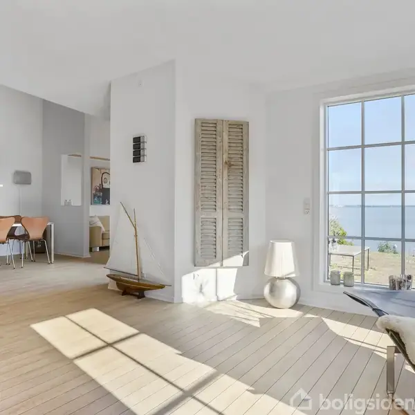 Stue med lyse trægulve og store vinduer viser havudsigt. Til venstre ses spisebord med stole og farverigt maleri. Væglampe og stående bådmodel i hjørnet.