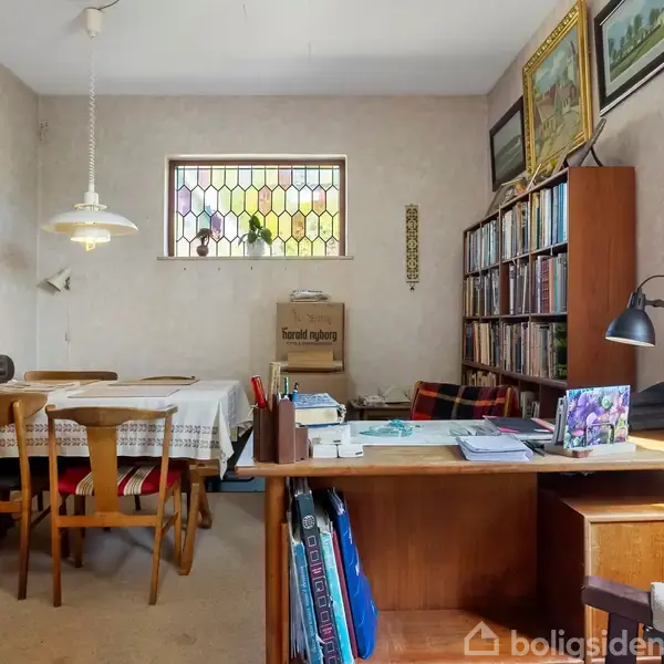 Et værelse med et spisebord til venstre dækket med en dug og omgivet af stole. En reol med bøger står i baggrunden. Et skrivebord med papirarbejde og en lampe findes til højre.
