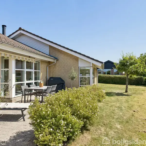 En bungalow med gul murstensfacade og store vinduer har en terrasse med spisebord og stole. Omgivet af en velplejet have med græsplæne og buske under en klar, blå himmel.