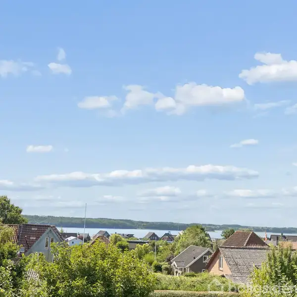 Huse med skrå tage, omgivet af frodig grønne træer og buske, med en udsigt over en fjord i baggrunden under en klar blå himmel med få skyer.