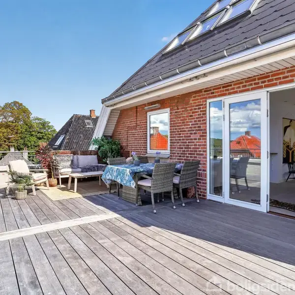 En træterrasse foran et murstenshus med åbne franske døre. Terrassebord med stole og plantepotter. Omgivet af andre hustage og grønne træer under en blå himmel.