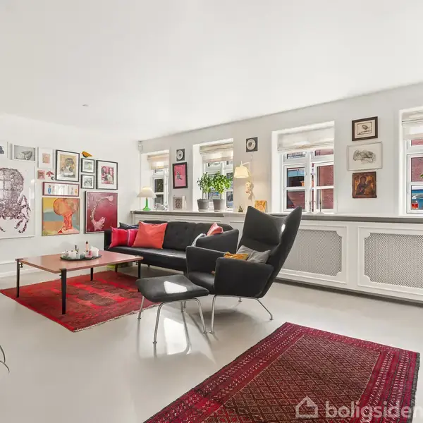 En stue med en sort sofa omkranset af kunst på væggene og planter i vindueskarmen. Rummet har lyst gulv, røde tæpper og store vinduer, der slipper naturligt lys ind.