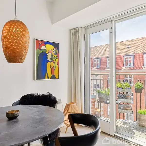 Et rundt spisebord med en enkelt skål står under en hængende, brun lampe. Et farverigt kunstværk hænger på væggen. Store vinduer med udsigt til en bygnings facade og altan med planter.