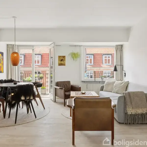 Stue med spisebord og stole dækket af fåreskind. En sofa og lænestol står ved siden af store vinduer med udsigt til røde hustage, omgivet af moderne dekorationer.