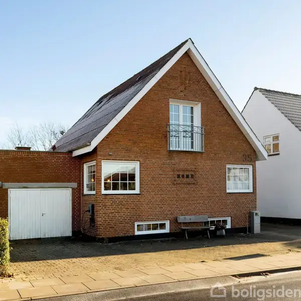 En murstensvilla med saddeltag står stille i et beboelsesområde. Foran er en garage og en bænk ved siden af hoveddøren. Sollys skinner på huset.
