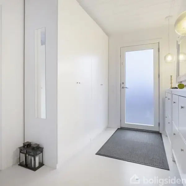 En lys entré med et hvidt skoskab til højre, en glasdør for enden, og en lanterne på gulvet til venstre. Der hænger en lampe ved siden af spejlet.