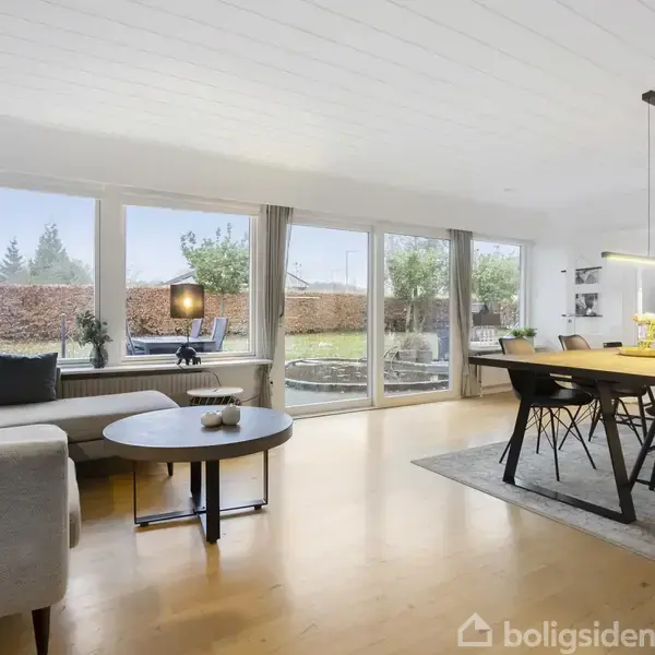 Stue med sofa og sofabord, oplyst af store vinduer. Et spisebord med stole står til højre, mens en lampe hænger over bordet. Udsigt til en have udenfor.