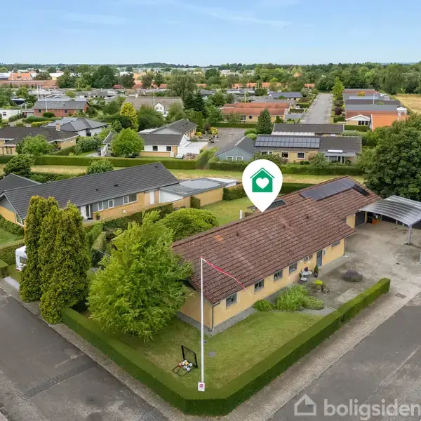 Et hus med rødt tegltag og velholdt have står på en hjørnegrund i et villakvarter. En carport er ved siden af huset, omgivet af hække og træer.