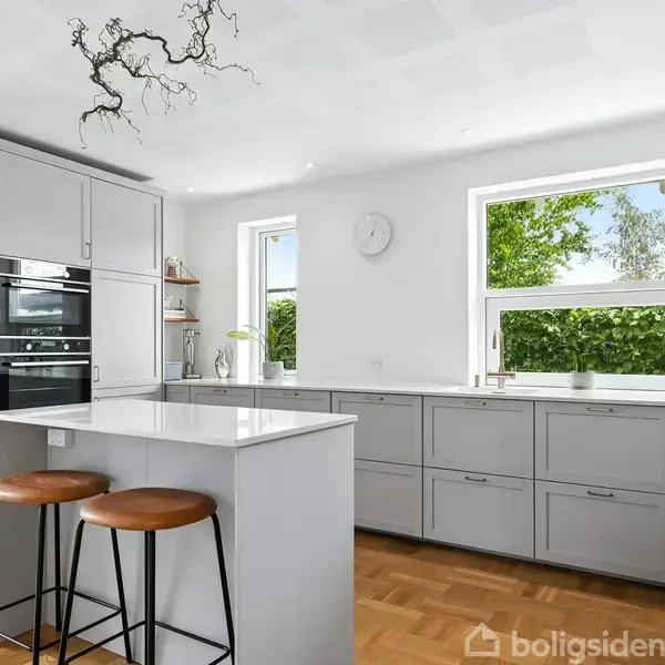 Køkken med grå skabe, to brune barstole ved en ø. Store vinduer viser grønne træer udenfor. En moderne ovn er indbygget, og en dekorativ lampe hænger fra loftet.