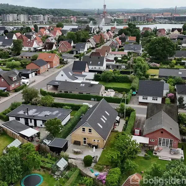 En bydel med rækkehuse og grønne haver ses oppefra. Husene står i rækker langs veje, med udsigt til en by og skovklædte bakker i baggrunden.