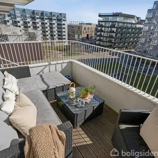 Terrassemøbler med grå puder står på en altan. Et bord med planter og drikkevarer er i centrum, omgivet af moderne lejlighedsbygninger i en urban bebyggelse.