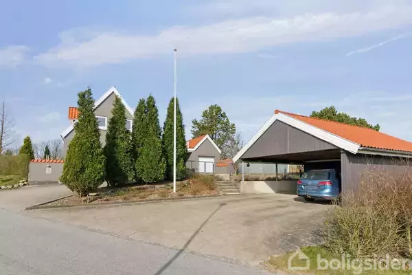 Hus med grå facader og orange tag står på en solrig dag. En blå bil holder parkeret i en carport. Omgivet af høje buske og asfaltvej.
