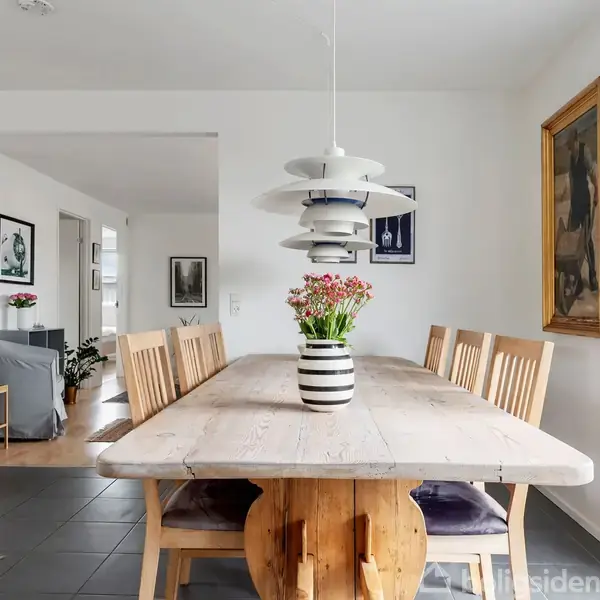 Et langt træbord med seks stole omgivet af lysere vægdekorationer. En stribet vase med blomster står på bordet, og en moderne lampe hænger over det. I baggrunden ses et sammenhængende opholdsrum.