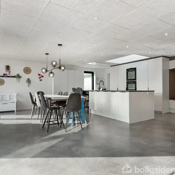 Et moderne køkken med et stort, hvidt køkkenbord og sorte stole ved spisebordet. Rummet har betonagtigt gulv, hvide vægge og loft, samt minimalistisk indretning med en lampe over bordet.